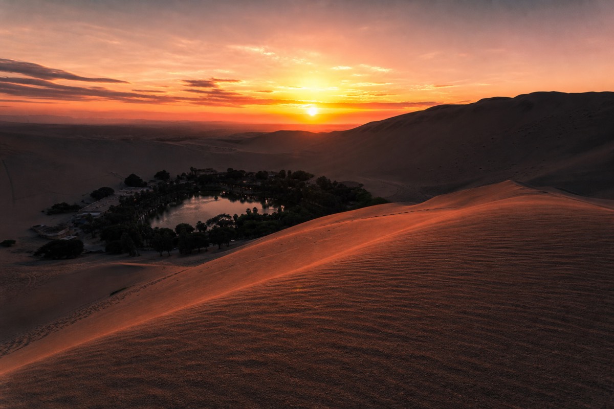 The small oasis town of Huacachina lit up at dusk with its lagoon reflecting the surrounding dune-top buildings